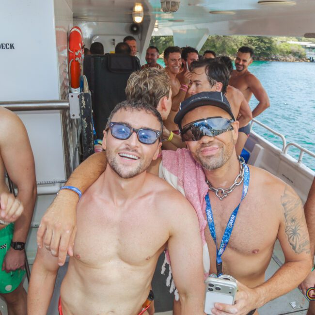 A group of men in swimwear smile and pose together on a boat. Two men in front, one with sunglasses and another with a cap and lanyard, have their arms around each other. The ocean and coastline are visible in the background.