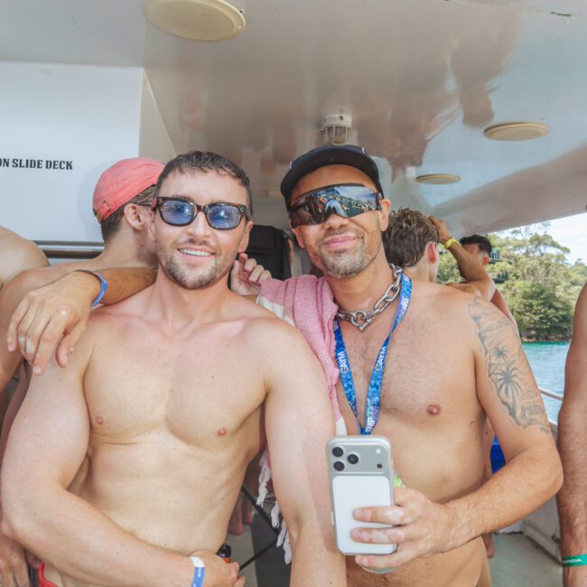Two men in swimwear and sunglasses smile and pose for a photo on a boat. One man holds a phone, and both are wearing lanyards. Other people and greenery are visible in the background.