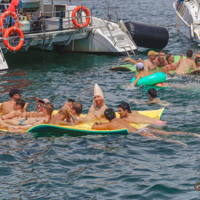 A group of men in swimsuits relax and socialize on floating mats in the water near several boats under sunny weather. Some people are wearing hats, and the atmosphere appears festive and lively.