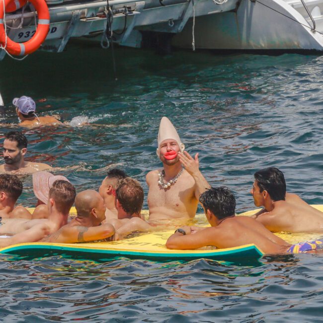 A group of men relax on a yellow floating mat in the water near a boat; one man in the center wears a clown nose, white clown hat, and makes a peace sign gesture.