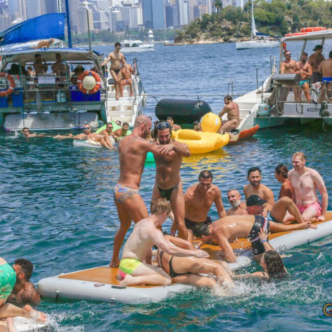 A group of people in swimsuits play and relax on floating mats and boats on a sunny day, with a city skyline and trees in the background. Some are splashing in the water, while others watch and laugh.