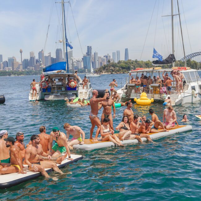 A lively group of people in swimwear enjoy a floating party on the water near boats, with a city skyline in the background. Some people stand and pose on an inflatable platform, while others relax and swim nearby.