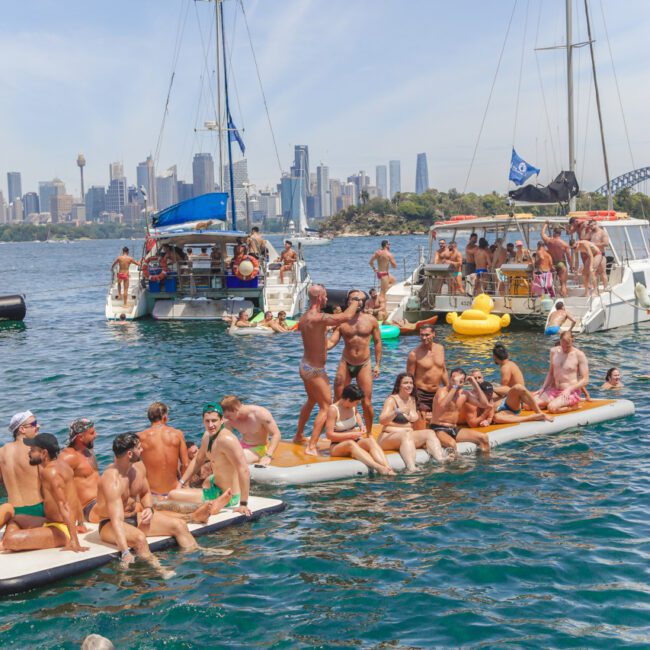 A group of people in swimwear are gathered on floating platforms and boats in a harbor, enjoying a sunny day. The city skyline and Sydney Harbour Bridge are visible in the background.