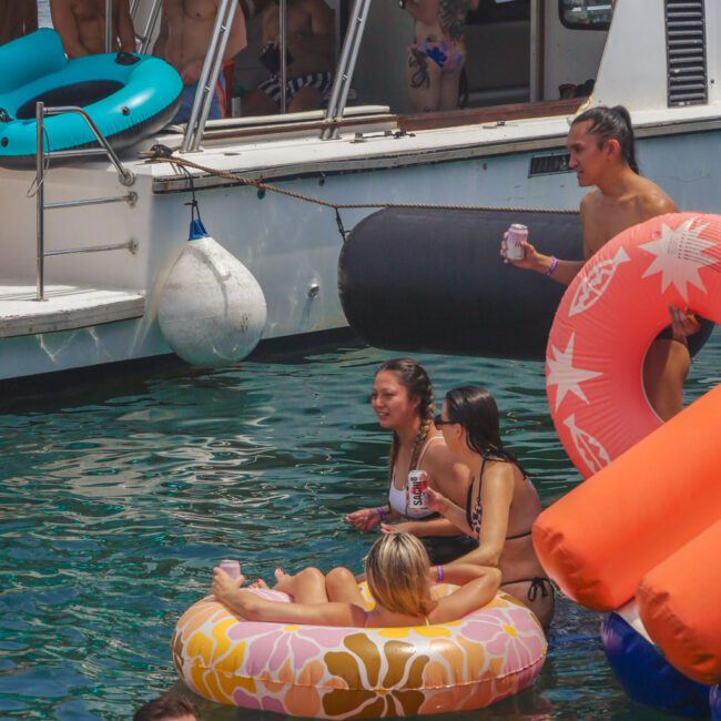 A group of young adults at a lake party, some on a boat and others relaxing on colorful inflatable tubes in the water, laughing and holding drinks on a sunny day.