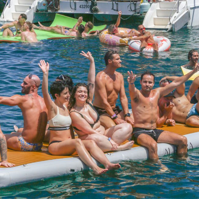A group of people in swimwear sit and wave on an inflatable float in the ocean, with others relaxing and socializing on a nearby boat under sunny weather.