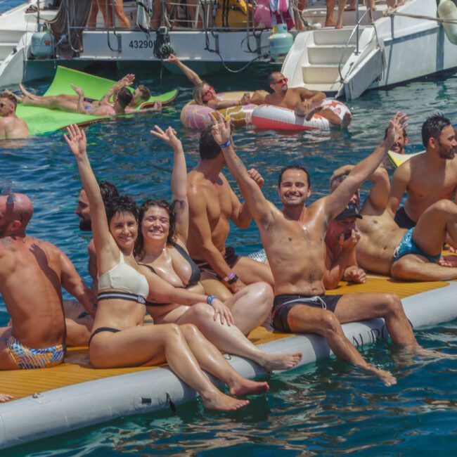 People in swimsuits relax and smile on a floating inflatable in the sea, with others swimming and lounging nearby and boats in the background. The scene is lively and joyful on a sunny day.