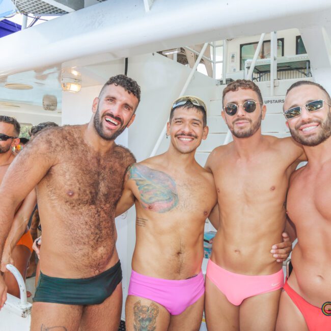 Four men in colorful swimwear smile and pose together on a boat. They are enjoying a sunny day, with other people and tropical scenery visible in the background. The atmosphere is festive and relaxed.