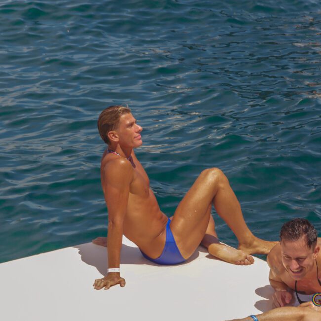 Two people in swimsuits relax on a white platform by the ocean, with one sitting and looking at the water while the other lies down and smiles.