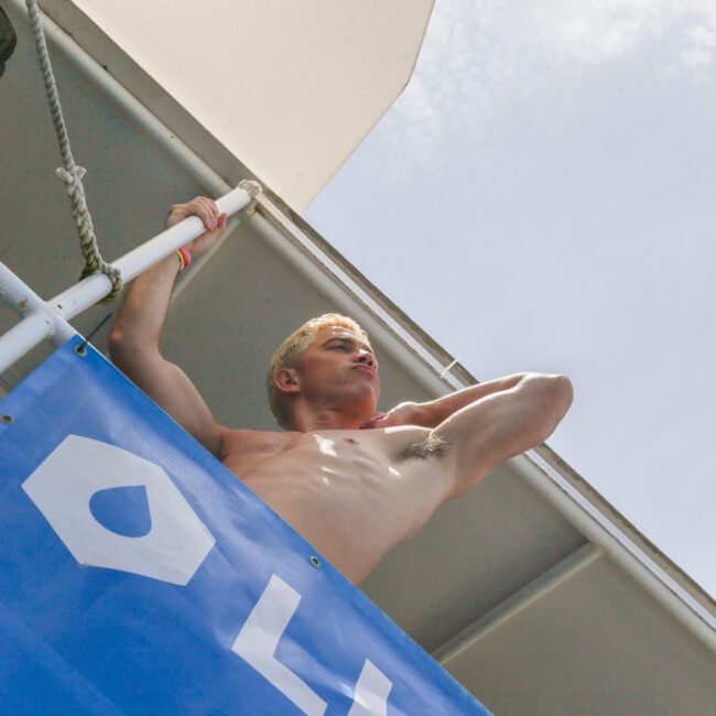 A shirtless person stands on a balcony under a white canopy, looking out with one hand shading their eyes. A blue banner hangs from the railing. The sky is bright and partly cloudy.