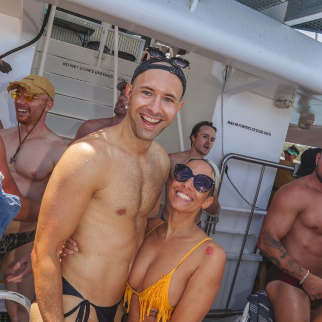 Two people in swimwear smile and pose together on a boat, surrounded by other people also in swimsuits. The atmosphere is cheerful and sunny, with a party vibe. Signs and boat equipment are visible in the background.