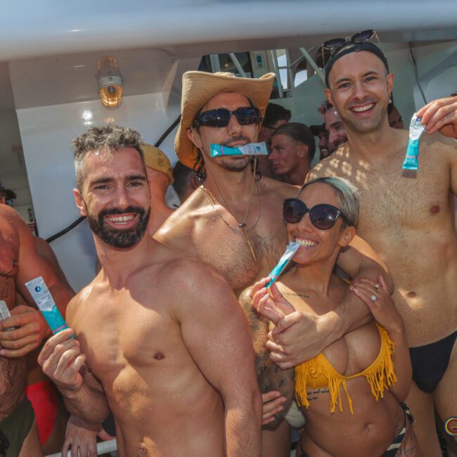 A group of smiling people in swimwear pose together at a sunny outdoor party, holding blue packets. Most are shirtless; one person wears a yellow bikini and sunglasses, another wears a cowboy hat. The atmosphere is festive and lively.