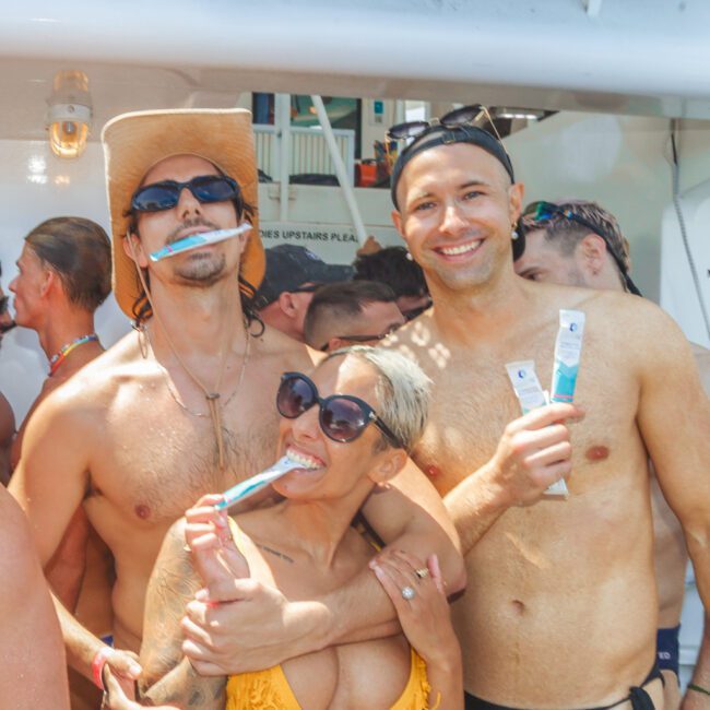 Three people in swimsuits smile and pose together at a crowded outdoor event, each holding or using a toothbrush. The group looks happy, with two men wearing hats and a woman in a yellow bikini and sunglasses.