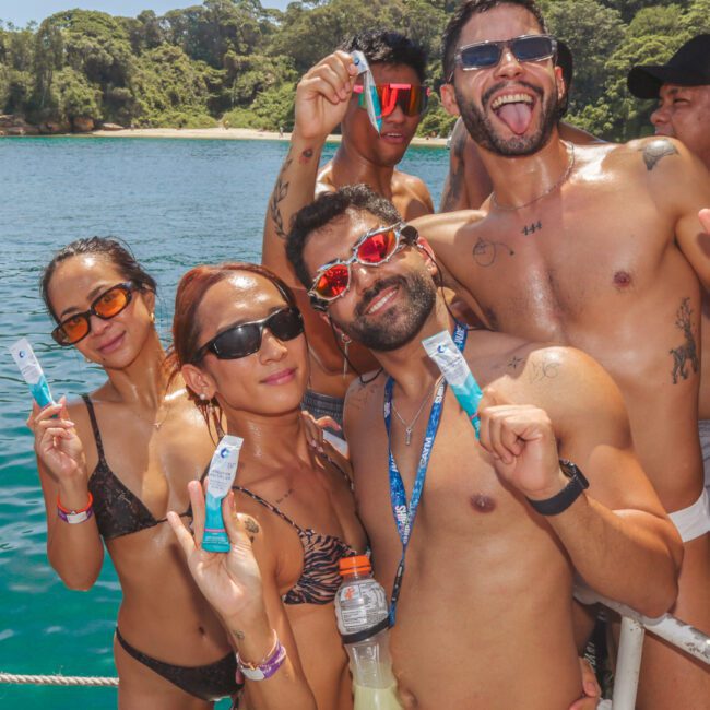 A group of people in swimwear and sunglasses smile and pose on a boat, holding up tubes of sunscreen, with blue water and lush green trees in the background on a sunny day.
