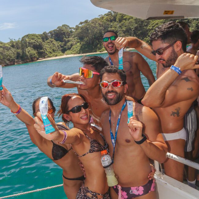 A group of people in swimsuits smile and pose with sunscreen packets on a boat, surrounded by blue water and lush greenery in the background.