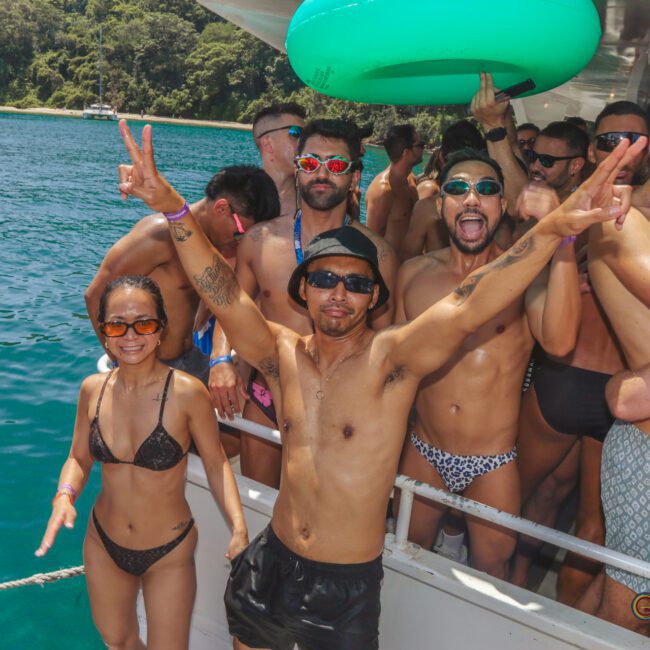 A group of people in swimwear are partying on a boat in sunny weather. Some are posing and smiling, with green trees and blue water in the background. One person holds up peace signs.