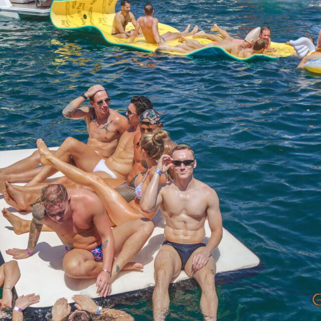 A group of people in swimwear relax and pose on floating mats and inflatables in the water on a sunny day. Some are smiling and saluting; others lounge or interact. The atmosphere is lively and festive.
