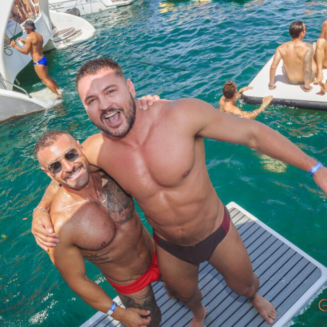 Two men in swimwear smile and pose together on a floating platform, surrounded by others enjoying a sunny day on the water with boats and swimmers in the background.