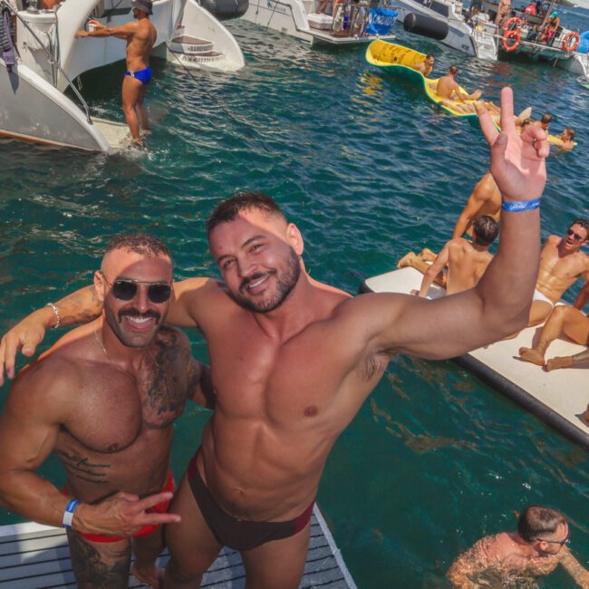 Two men in swimwear smile and pose on a boat platform, surrounded by others enjoying a sunny party on boats and in the water. One man holds up a peace sign. The scene is lively and festive.