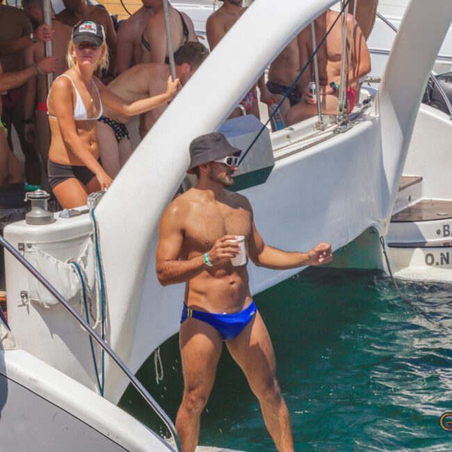 A group of people in swimsuits enjoy a sunny day on a boat party, with one person in blue swim briefs and a black hat standing near the edge of the boat holding a drink. Other boats and partygoers are in the background.