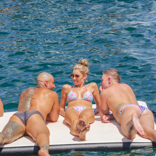 Three adults in swimwear lie on their stomachs on a floating platform in the water, facing each other and smiling under the sun. The water is bright blue and sparkles in the background.