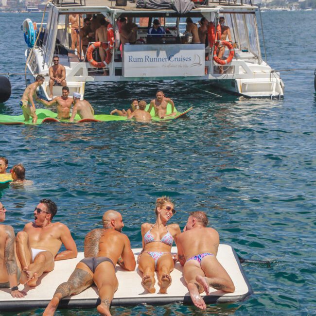 People relax on a floating platform and swim in the water near a Rum Runner Cruises boat. The weather is sunny, and the scene is lively with groups enjoying the ocean and city skyline in the background.