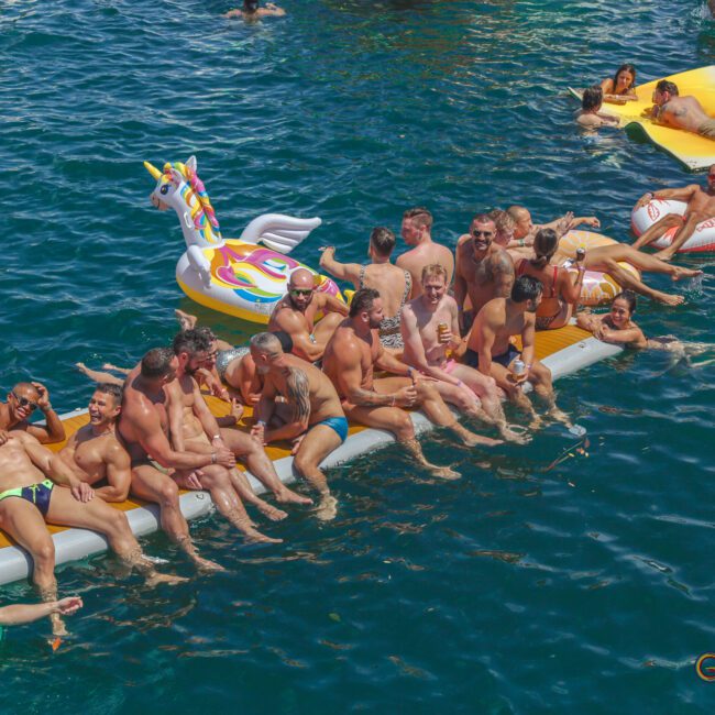 A group of men in swimwear relax and pose on inflatable rafts, including a unicorn float, in a vibrant blue body of water during a sunny day. Other people float nearby on various inflatables.