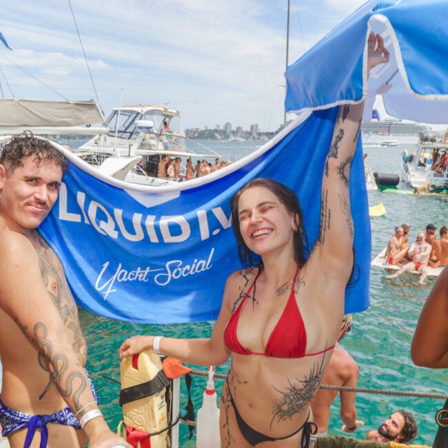 Two people in swimsuits hold a blue "LIQUID I.V. Yacht Social" banner, smiling on a boat amid a lively crowd. Other boats and people in swimsuits are visible on the water under a sunny sky.