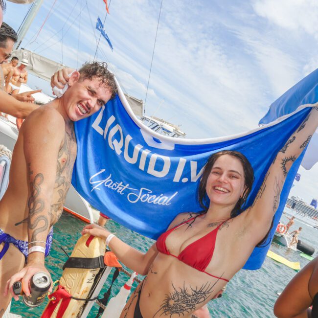Two people in swimwear smile and pose on a boat, holding up a blue towel reading "LIQUID I.V. Yacht Social;" other people and a city skyline are visible in the background over turquoise water.