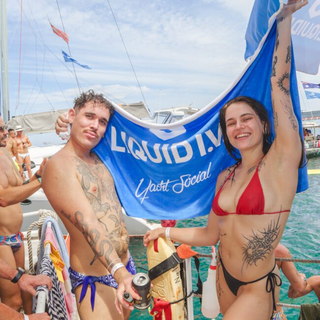 Two smiling people in swimsuits stand on a dock near yachts, holding up a blue "Liquid I.V. Yacht Social" towel. Others in swimwear are seen in the background, enjoying a sunny day by the water.