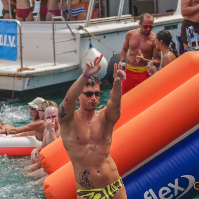 A man in patterned swim trunks dances with his arms up on a boat deck near an orange inflatable slide, while others relax and socialize in the water and on pool floats at a lively pool party.