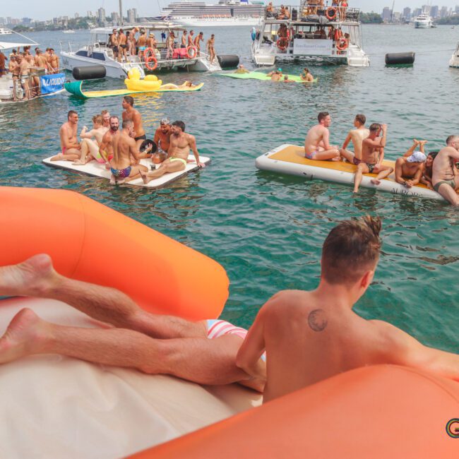 Groups of people in swimsuits relax on inflatables and boats in a crowded, lively water party. One man lounges in the foreground, looking out over the scene. The city skyline and yachts are visible in the background.