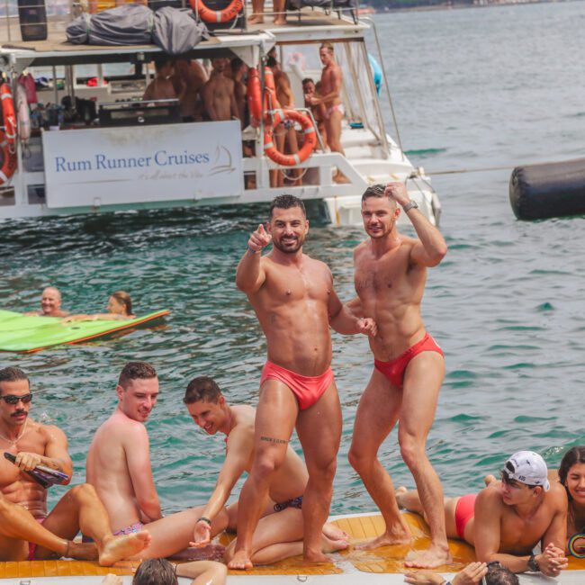 A group of people in swimsuits enjoy a lively boat party on the water. Two men in matching red swim briefs stand and pose on a floating platform, while others sit and relax nearby. A boat labeled "Rum Runner Cruises" is in the background.
