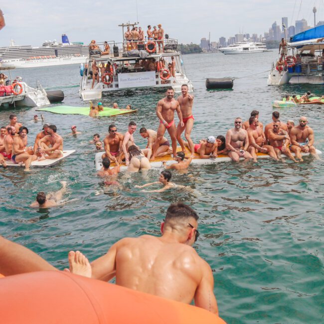 A lively group of people in swimsuits enjoy a party on boats and in the water near a city shoreline; some stand on floating platforms, others swim or sit, with city buildings visible in the background.