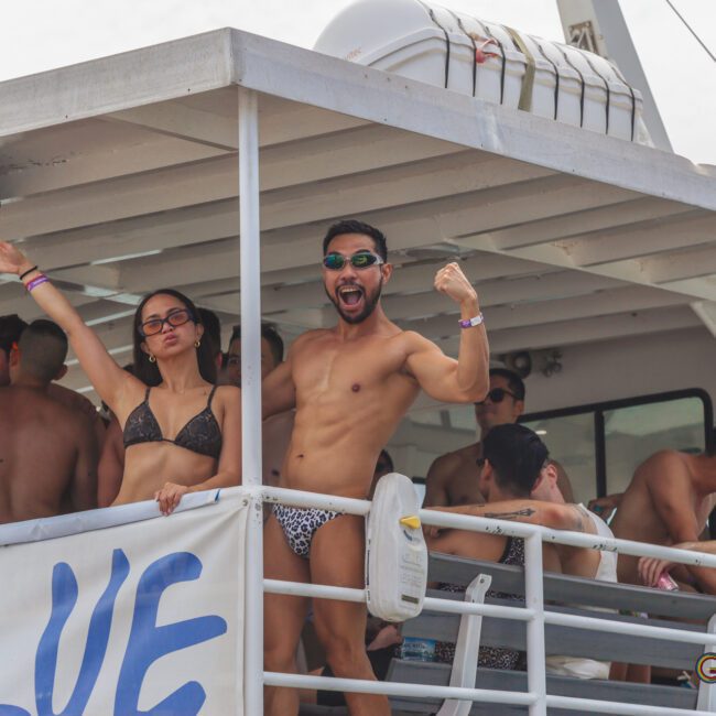 A group of people in swimwear enjoy a party on a boat. Two people at the front, one flexing and cheering, the other raising an arm and smiling, look at the camera. Others socialize in the background.