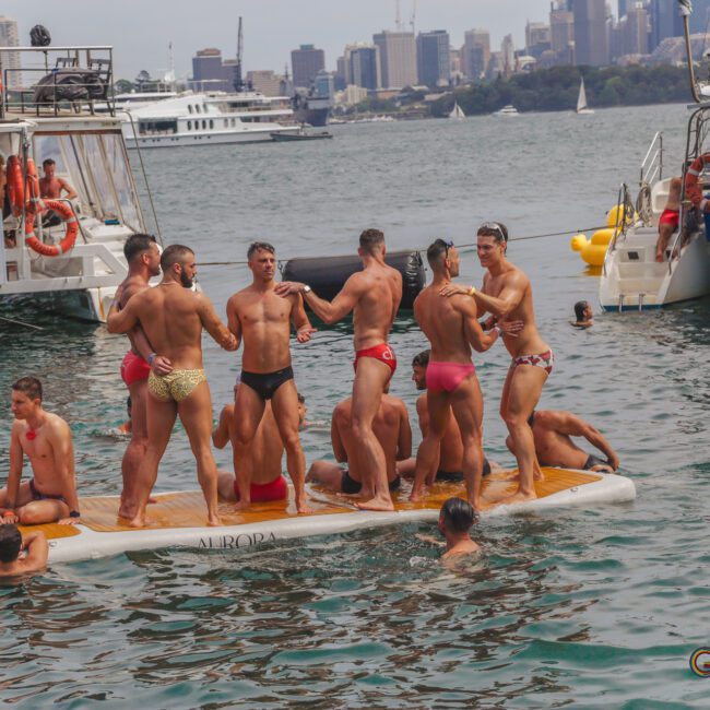 A group of men in colorful swimwear stand and socialize on a floating platform in a busy harbor, with boats and swimmers around them and a city skyline in the background.