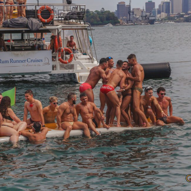 A group of men in swimwear stand and sit close together on an inflatable platform in the water near a party boat, with a city skyline visible in the background.