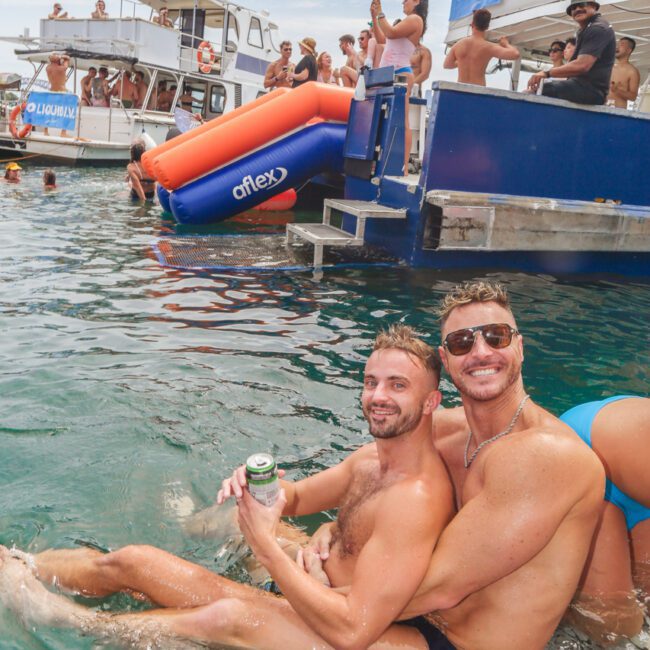 Two men in swim trunks sit on the edge of a boat in the water, smiling and embracing. Other people are on nearby boats and in the water, enjoying a lively, sunny party atmosphere.