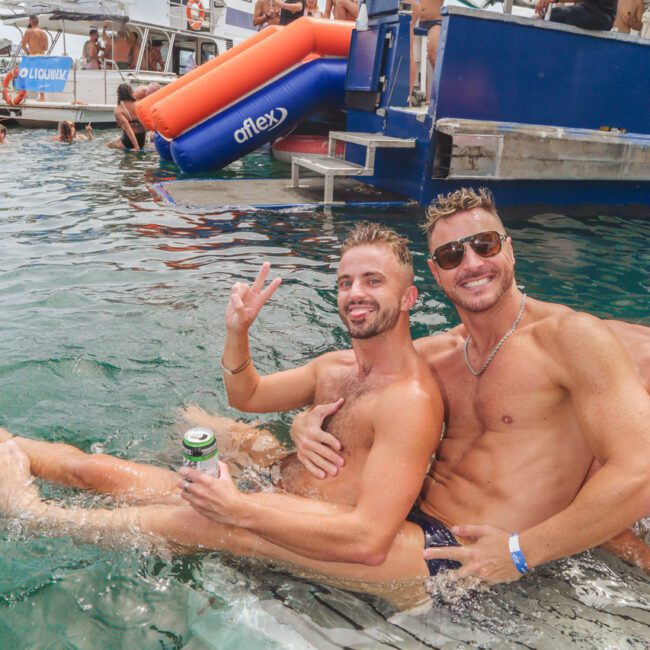 Two men sitting on a floating mat in the water, smiling at the camera; one holds up a peace sign, and people are partying on boats and a dock in the background.