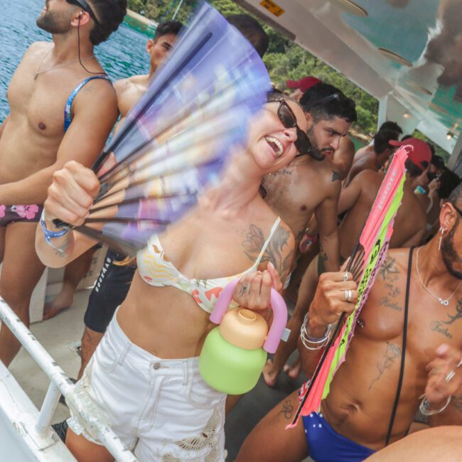A group of people in swimwear dance and smile on a boat, holding colorful fans and drinks. The sunny scene takes place by the water, with green trees visible in the background.