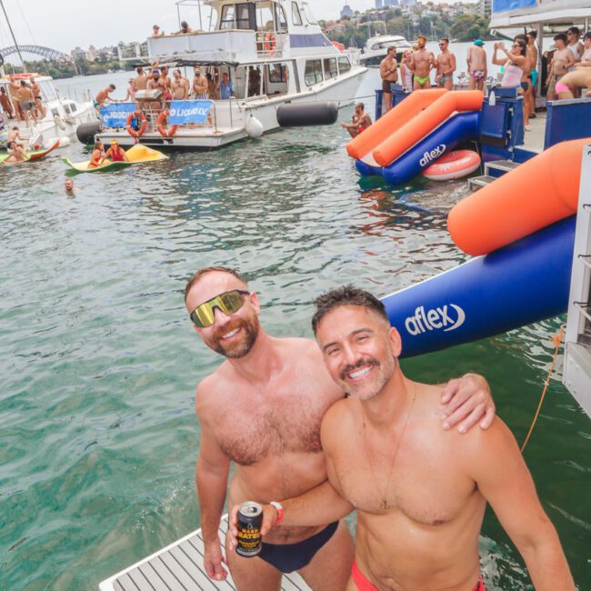 Two men in swimwear smile and pose on a floating dock at a lively boat party, holding drinks. People are swimming and relaxing on boats and inflatables in the background under a partly cloudy sky.