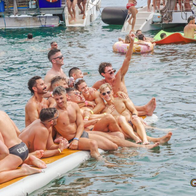 A group of men in swimwear sit closely together on an inflatable float in the water, smiling and posing for a photo. Other people are seen swimming and relaxing nearby, with boats in the background.