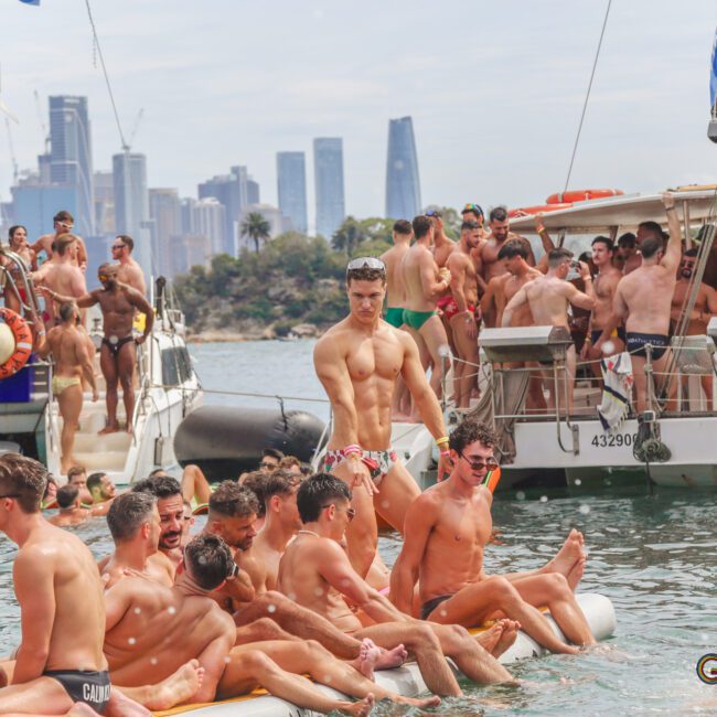 A large group of men in swimwear gather on boats and an inflatable raft in the water, with a city skyline in the background on a sunny day. The scene appears lively and festive.