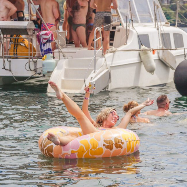 A woman in sunglasses lounges joyfully on a colorful inflatable ring in the water, holding a drink, with a group of people partying on a nearby boat in the background.