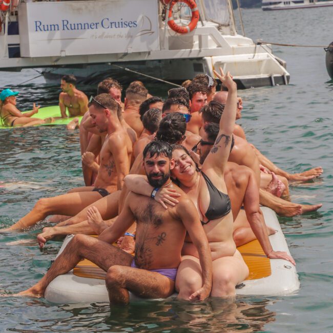 A group of people in swimwear sit closely together on a floating mat in the water near a Rum Runner Cruises boat, smiling and posing for photos during a sunny day.