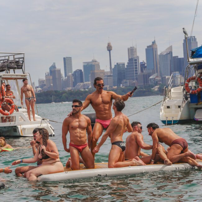 A group of people in swimwear enjoy themselves on a floating mat in the water, surrounded by boats, with a city skyline and the Sydney Opera House visible in the background.
