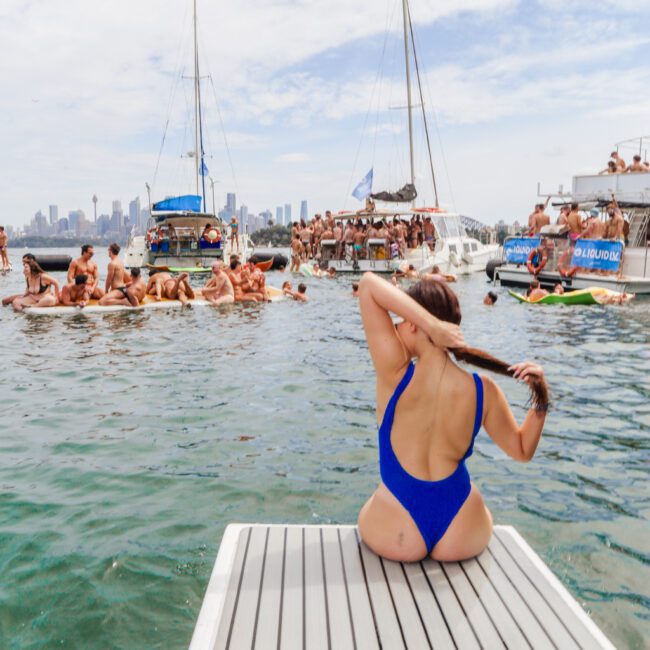 A woman in a blue swimsuit sits on a dock facing the water, with boats and groups of people partying and swimming in the background under a blue sky. The city skyline is visible in the distance.