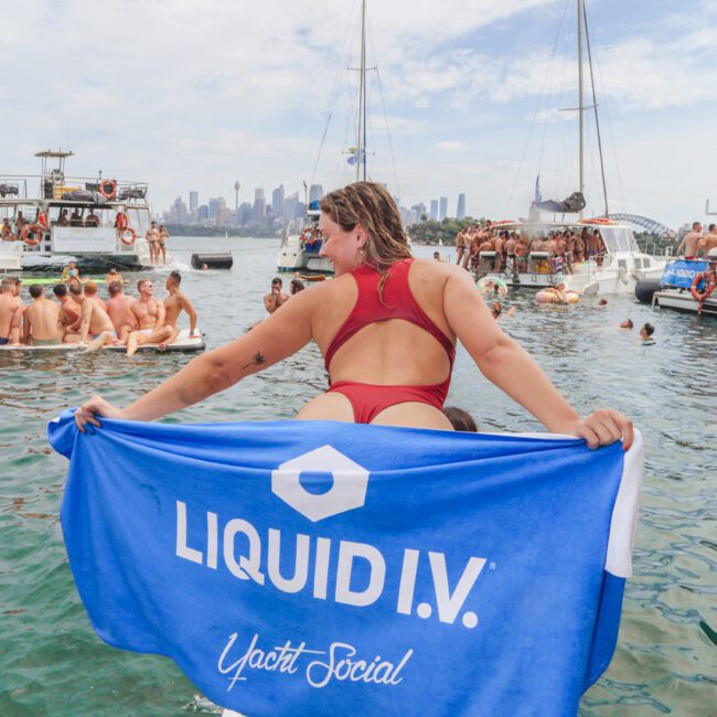 A person in a red swimsuit stands on a dock holding a blue "LIQUID I.V. Yacht Social" towel, with boats and a crowd of people enjoying a party on the water in the background. The city skyline is visible in the distance.