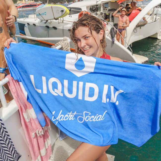 A smiling woman stands on a yacht holding a large blue towel that reads "LIQUID I.V. Yacht Social." People are relaxing on the yacht and another boat is visible in the background on the water.