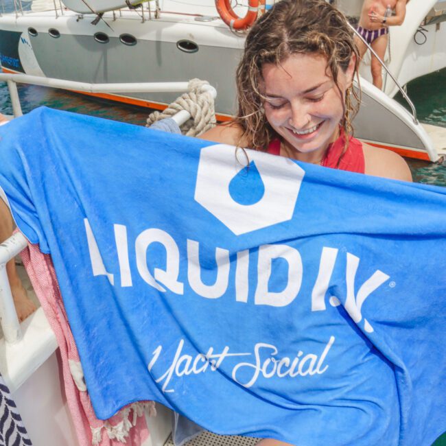 Smiling woman on a boat holds a blue "LIQUID I.V. Yacht Social" towel. In the background, a man on another boat raises his arms in celebration near kayaks and a sailboat.