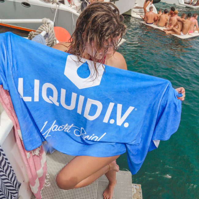 A person with wet hair holds a blue "LIQUID I.V. Yacht Social" towel on a boat, with groups of people partying on nearby boats and in the water under a sunny sky.